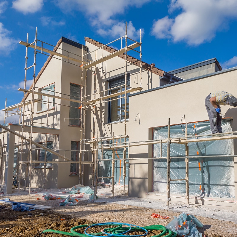 Maison neuve en construction avec échafaudage et façade beige finie. Un ouvrier travaille sur une fenêtre sous ciel bleu.