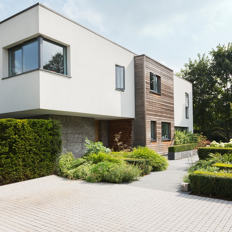 Maison moderne à l'architecture épurée, façade blanche et bois, avec jardin paysager verdoyant et allée pavée en damier.