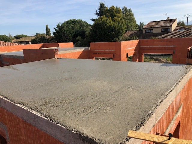 Dalle en béton fraîchement coulée sur les murs en briques rouges d'une maison en construction, sous un ciel bleu dégagé.