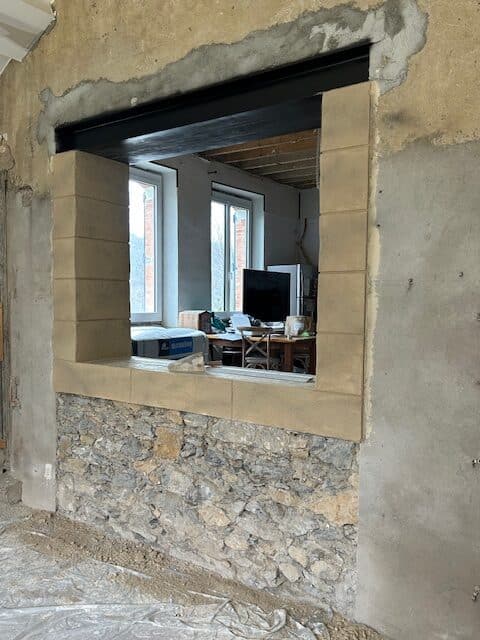 Mur en rénovation avec ouverture. Poutre métallique noire et blocs de pierre encadrent l'ouverture sur une base de maçonnerie brute.